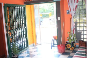 Una habitación con piso a cuadros, una puerta verde con celosía metálica, una ventana con cortina y una planta en maceta.