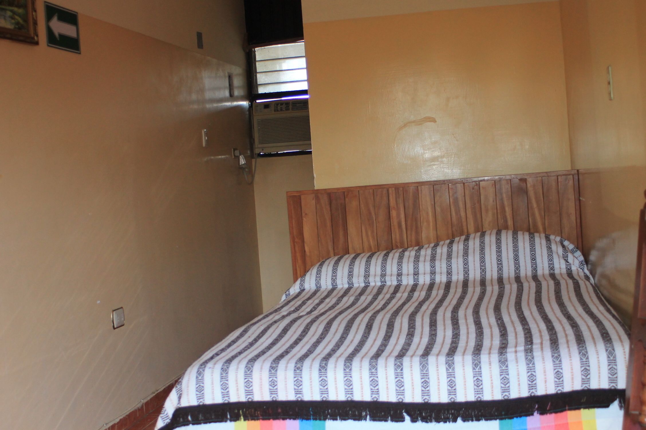 A room with a bed, a wooden headboard, a wall-mounted air conditioning unit, and a door with an arrow sign.