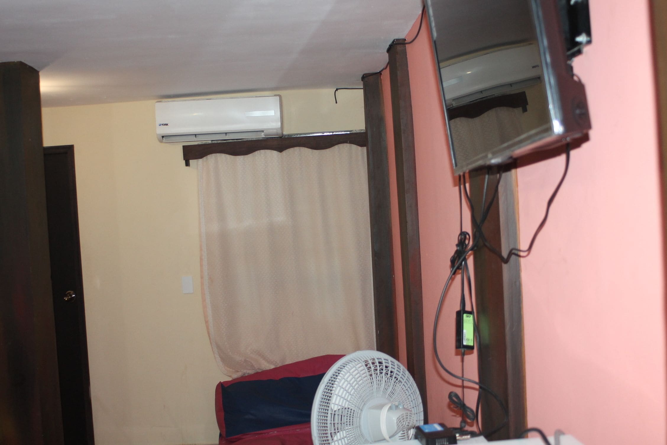 A room with a ceiling fan, an air conditioning unit, a television, and a bed with a red blanket.
