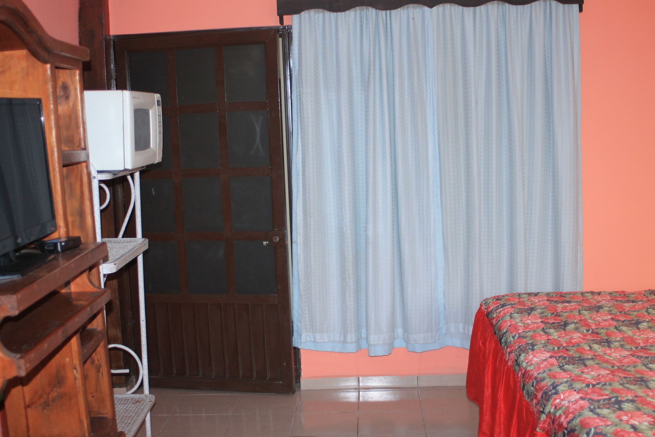 A room with a bed, a television on a wooden stand, a door with a glass panel, and a curtain.