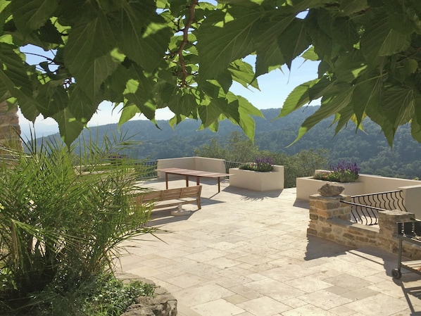 An outdoor patio with a table and chairs, surrounded by potted plants and overlooking a mountainous landscape.