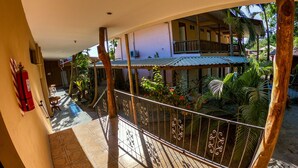 A covered balcony with a tiled floor, a fire extinguisher, and a view of a building with a balcony and plants.