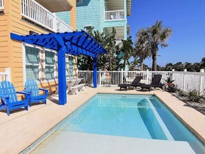 A swimming pool with blue chairs and a pergola, surrounded by a white fence and palm trees. - Sol Mates VW61 (Port Aransas)