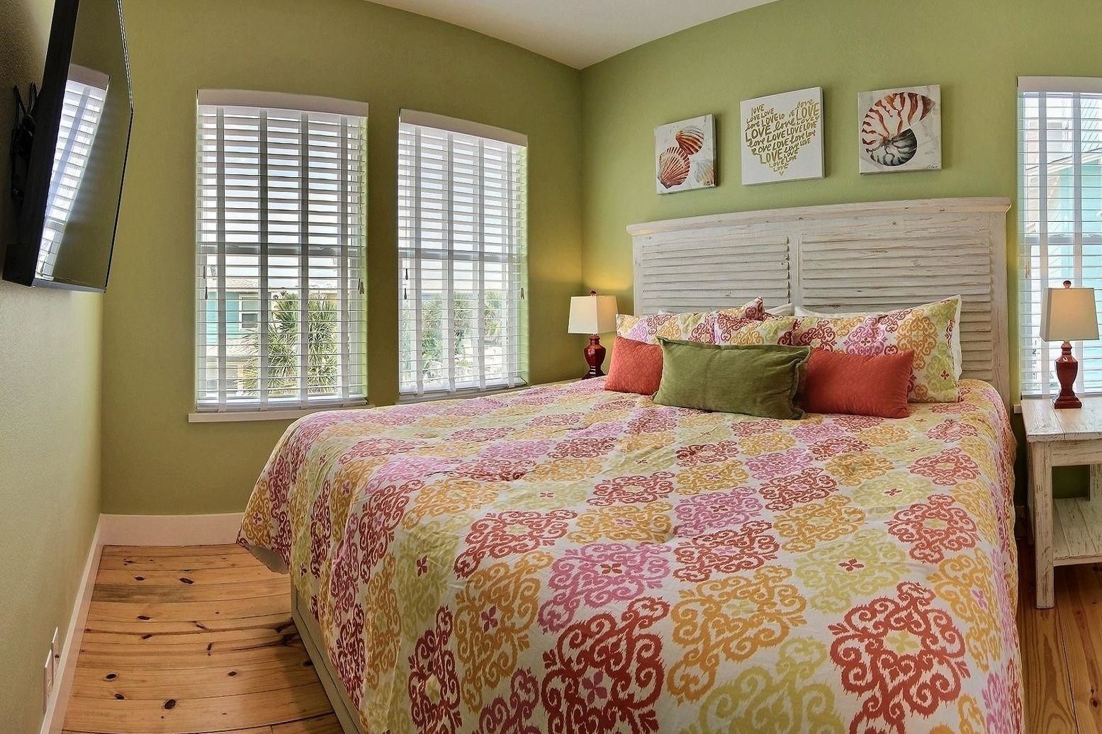 Un dormitorio con cama, dos ventanas con persianas, una mesita de noche con lámpara y decoración en la pared.