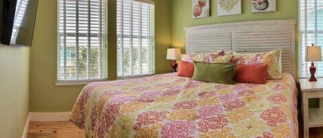 Un dormitorio con cama, dos ventanas con persianas, una mesita de noche con lámpara y decoración en la pared.