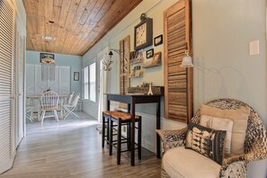 A room with a wooden ceiling, a dining area with a table and chairs, a bar area with a shelf, and a patterned armchair.