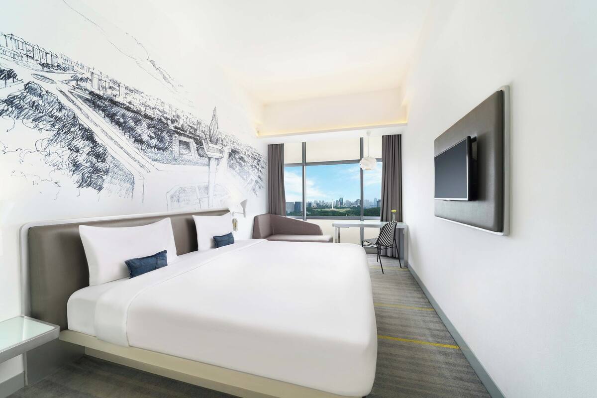 A modern hotel room with a large bed, a desk, and a chair. There is a large window with a view of the city.