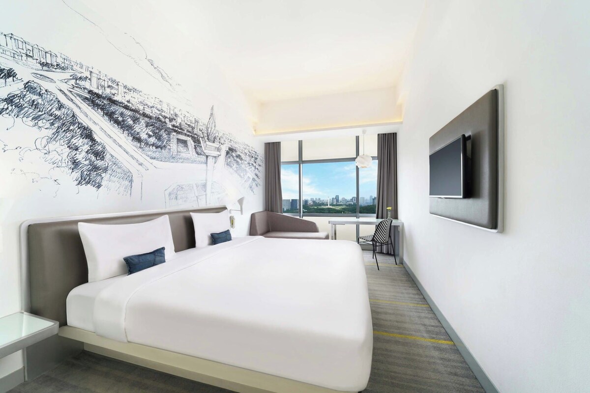 A hotel room with a large bed, a desk, a chair, a TV, and a cityscape view.