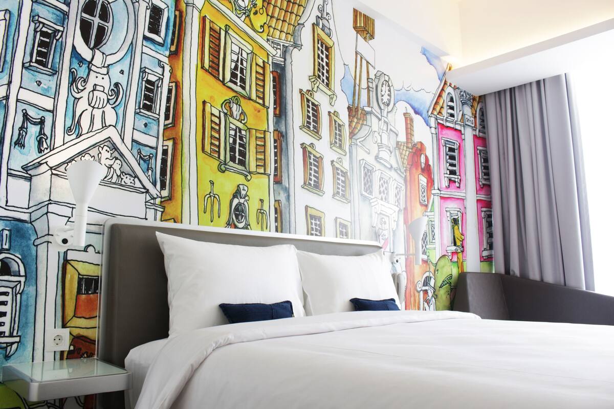 A hotel room with a bed, a nightstand, and a colorful mural on the wall.
