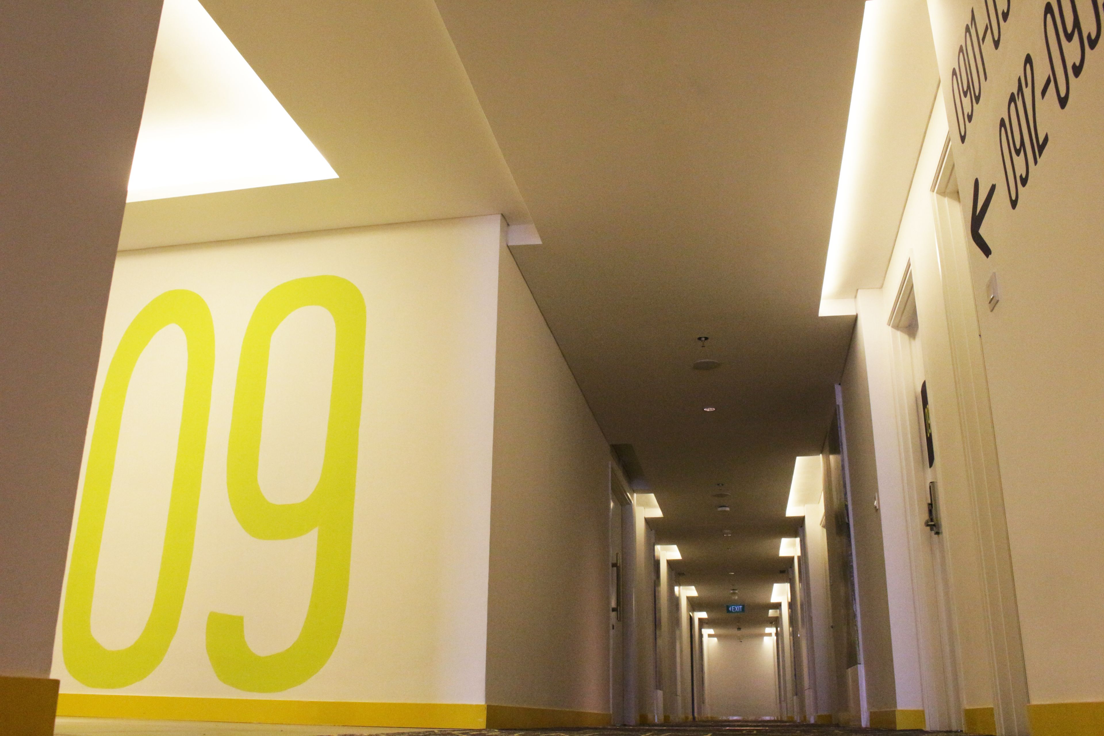 Un pasillo de hotel con un cartel grande que indica '09' a la izquierda, señalando el noveno piso. El pasillo está bien iluminado con luces del techo, y hay puertas a ambos lados que dan a las habitaciones.