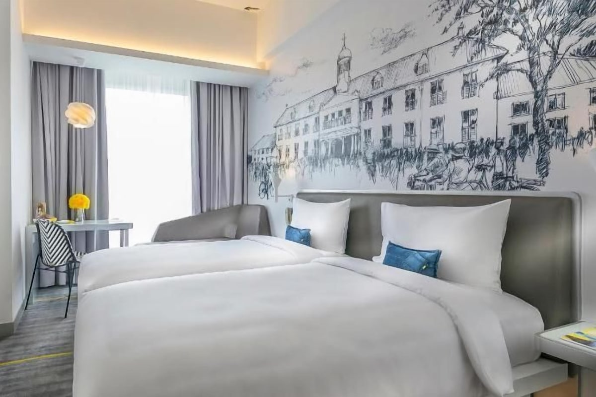 A hotel room with a large bed, a desk with a chair, a window with curtains, and a wall mural.