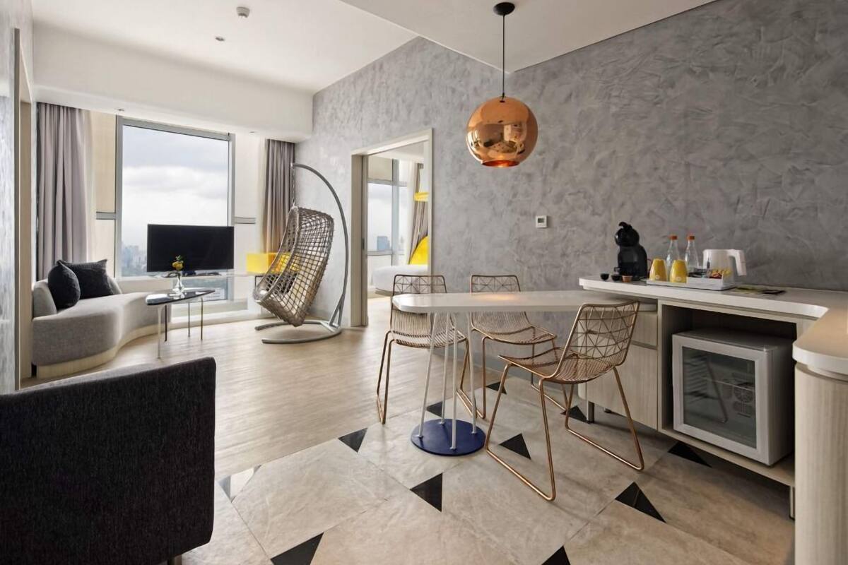 A modern living room with a sofa, a dining table with chairs, a kitchen area with a sink and counter, and a large window providing a view.