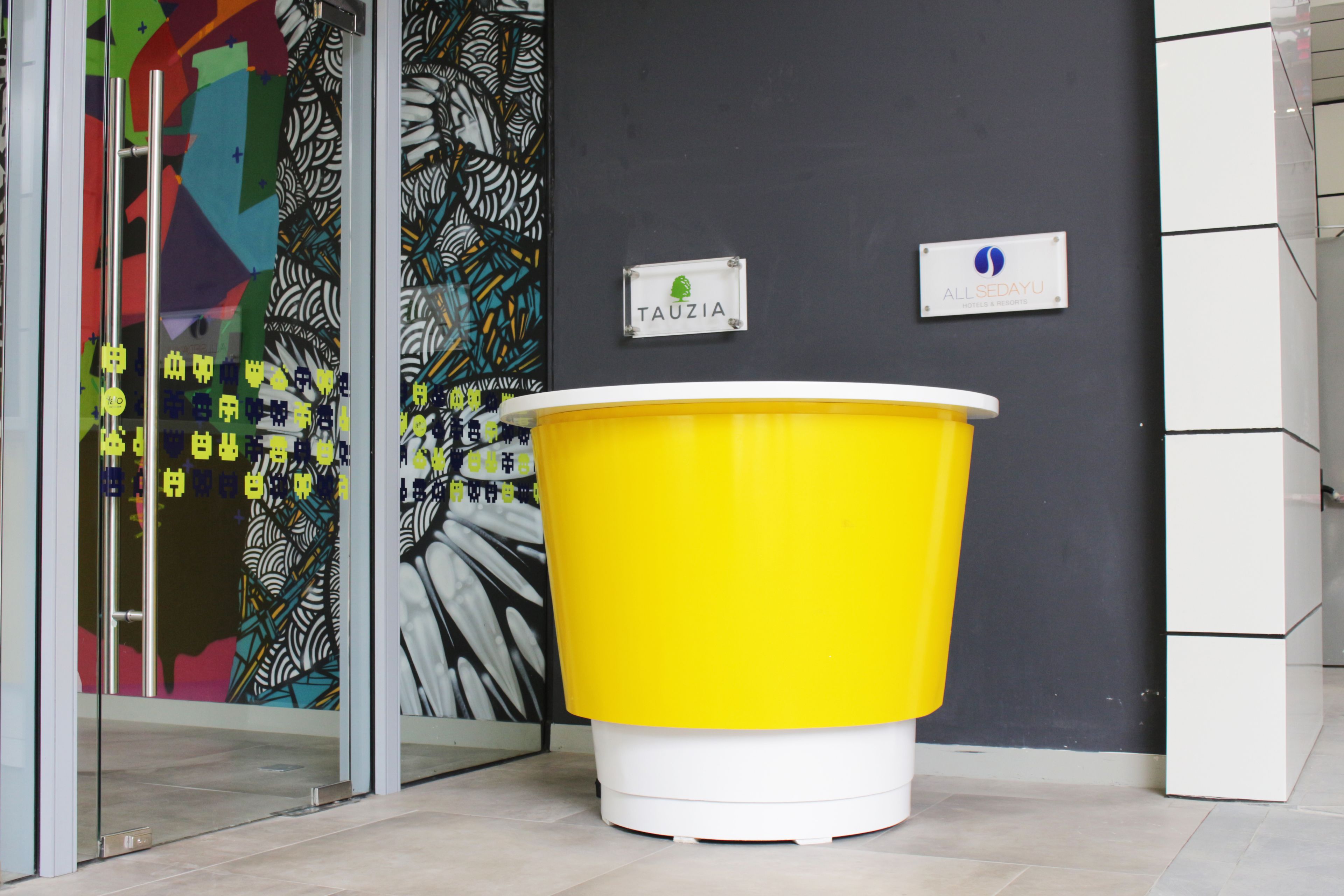 A reception area with a yellow trash bin, a glass door with colorful artwork, and two signs on the wall.