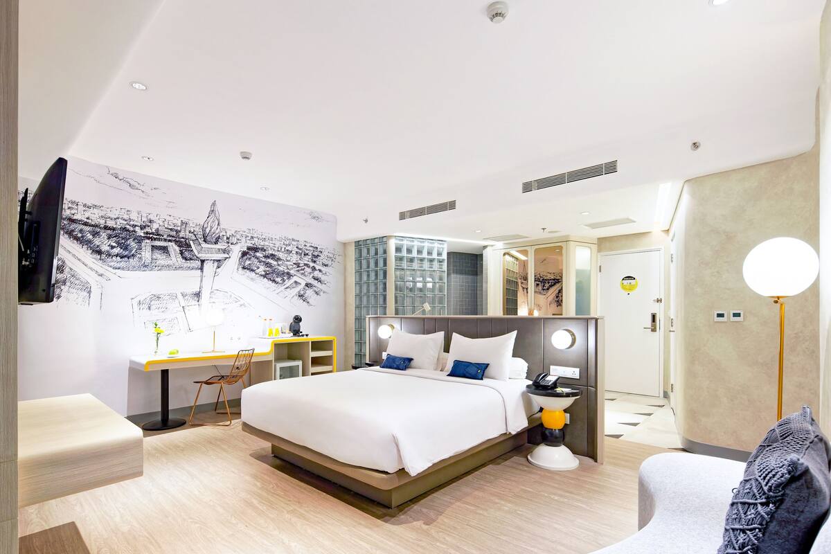 A modern hotel room with a large bed, a desk, a chair, and a sofa.