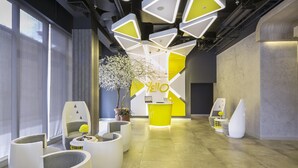 Un vestíbulo moderno con un mostrador de recepción amarillo, zonas de estar blancas y un amplio ventanal que proporciona luz natural.