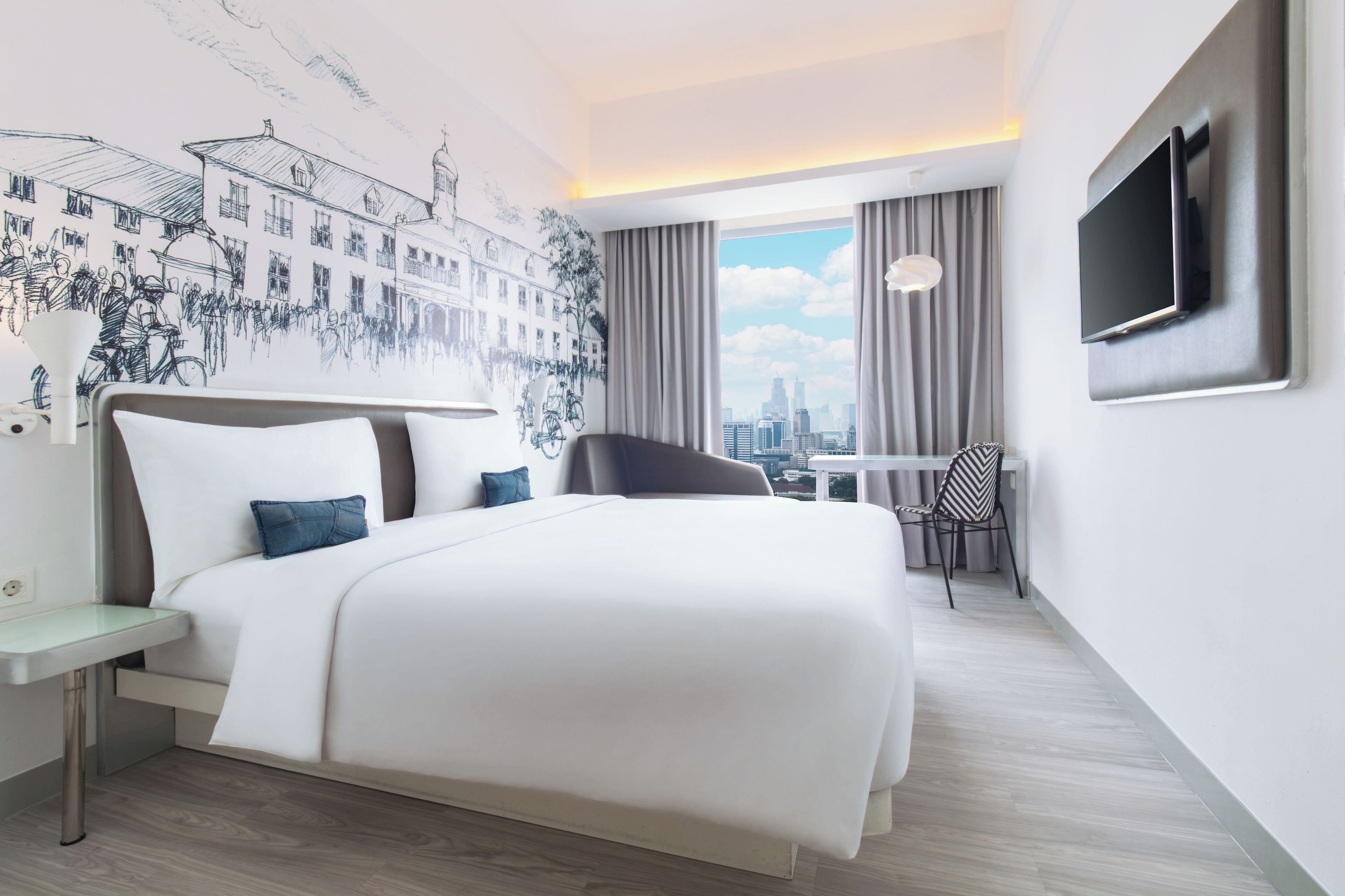 A hotel room with a large bed, a desk, a chair, a TV, and a cityscape view.