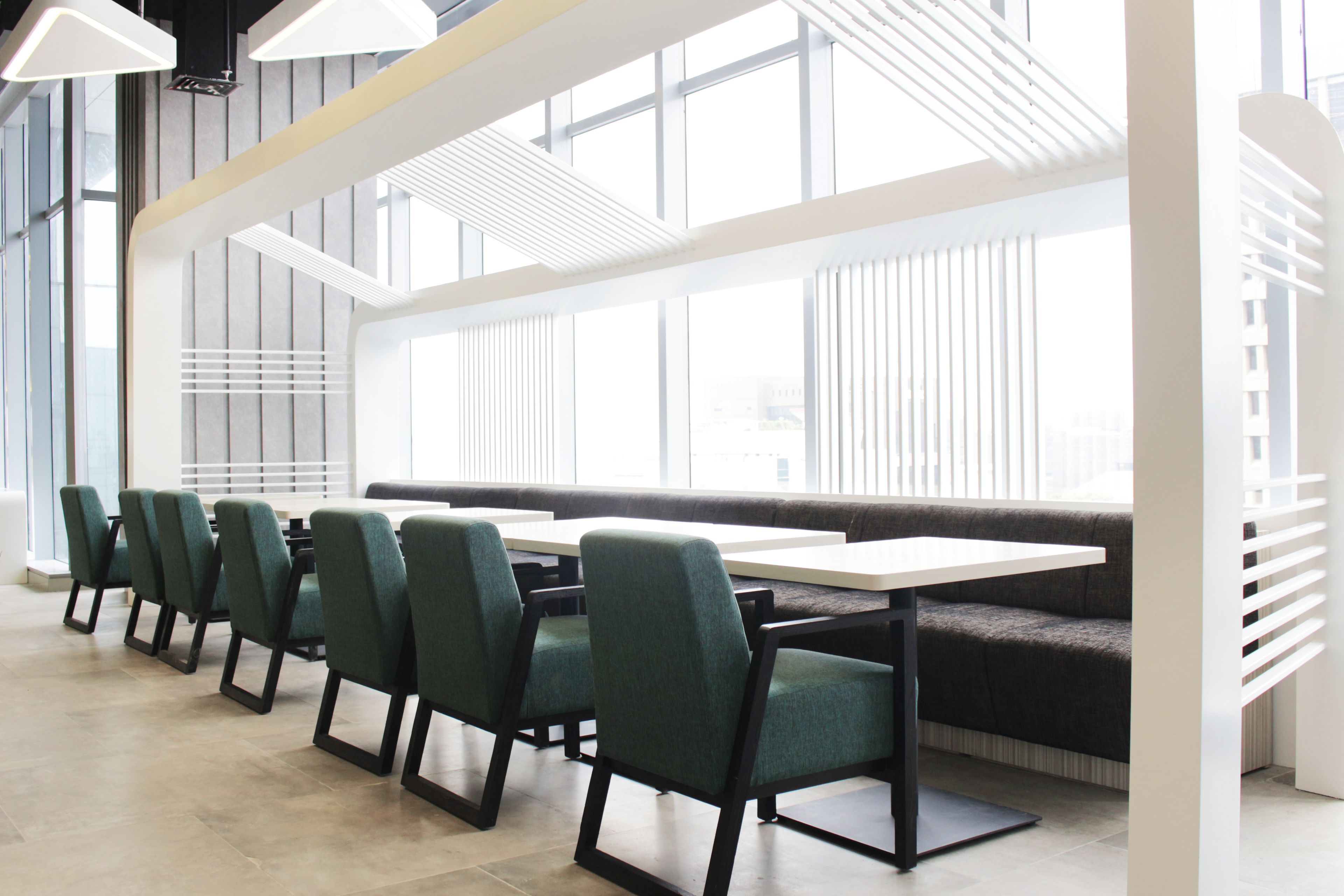 A modern dining area with a long table and chairs, large windows, and a high ceiling with lighting fixtures.