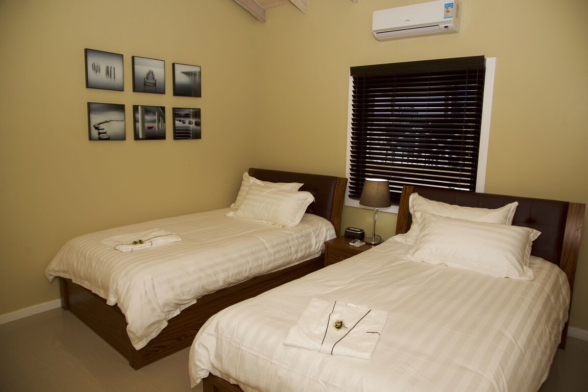 Une chambre d’hôtel avec deux lits, une fenêtre avec des stores, un climatiseur fixé au mur et six tableaux encadrés accrochés au mur.