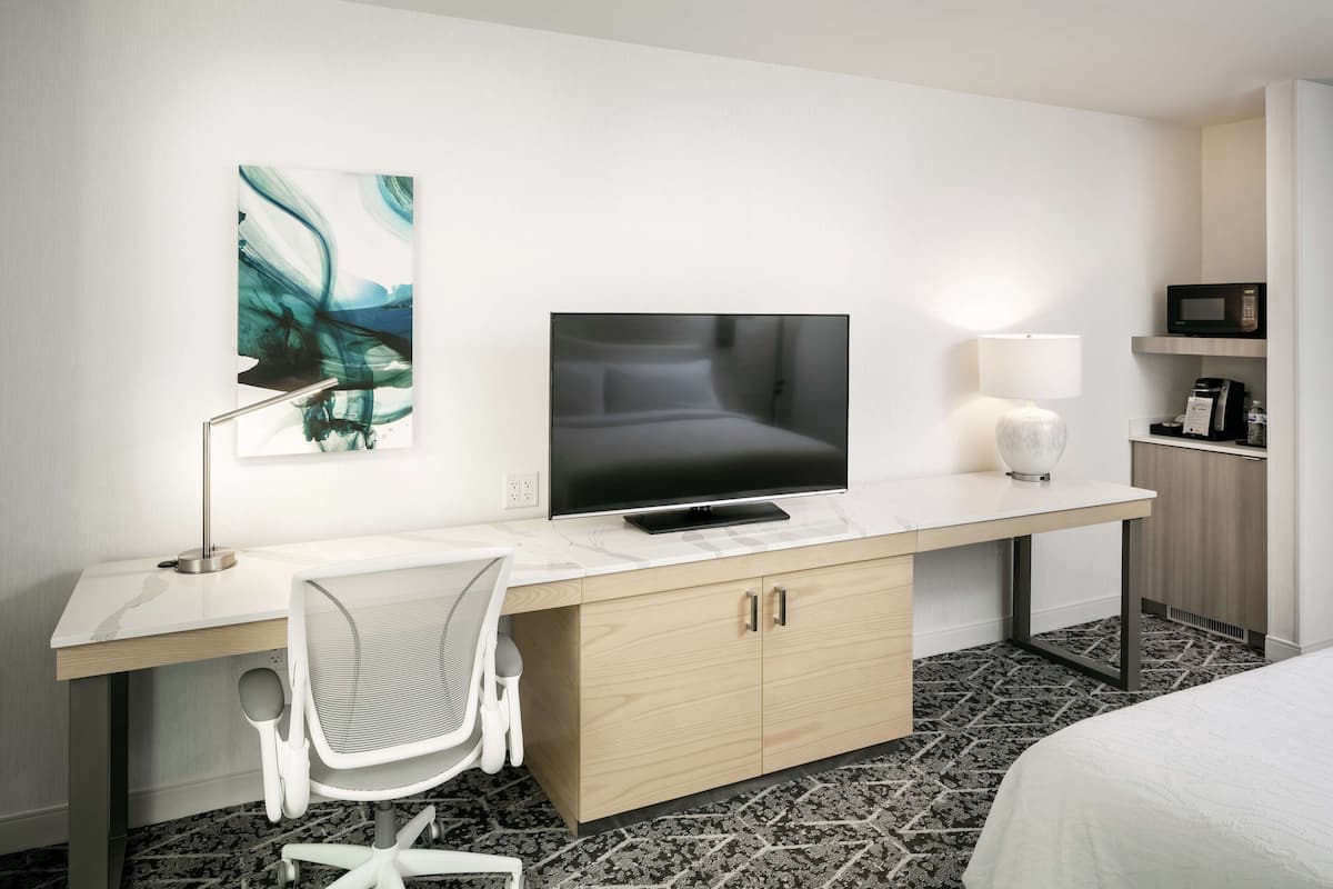 A hotel room with a desk, a television, a chair, a bed, and a microwave.