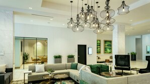 A modern hotel lobby with a sectional sofa, green armchairs, and a TV. There are multiple hanging light fixtures and potted plants. - Hilton Garden Inn Seattle Bellevue Downtown, WA (Bellevue)