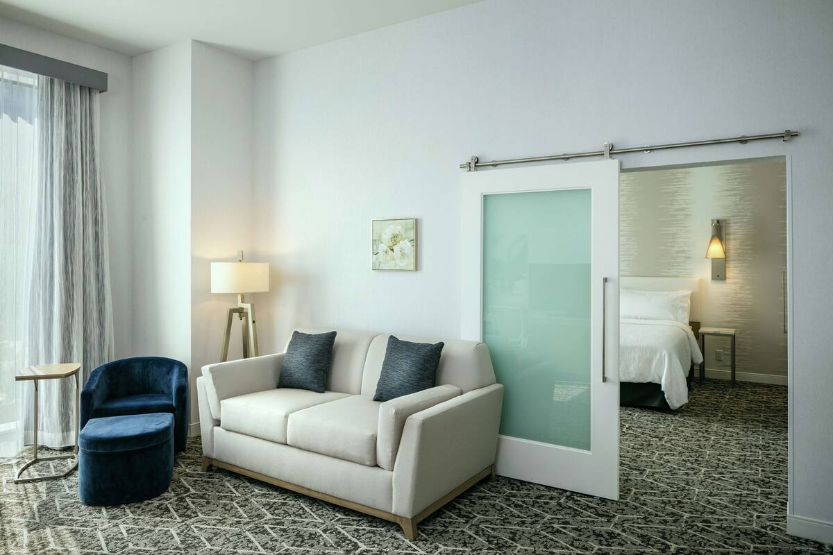 A hotel room with a sofa, a chair, a bed, a lamp, and a painting on the wall.