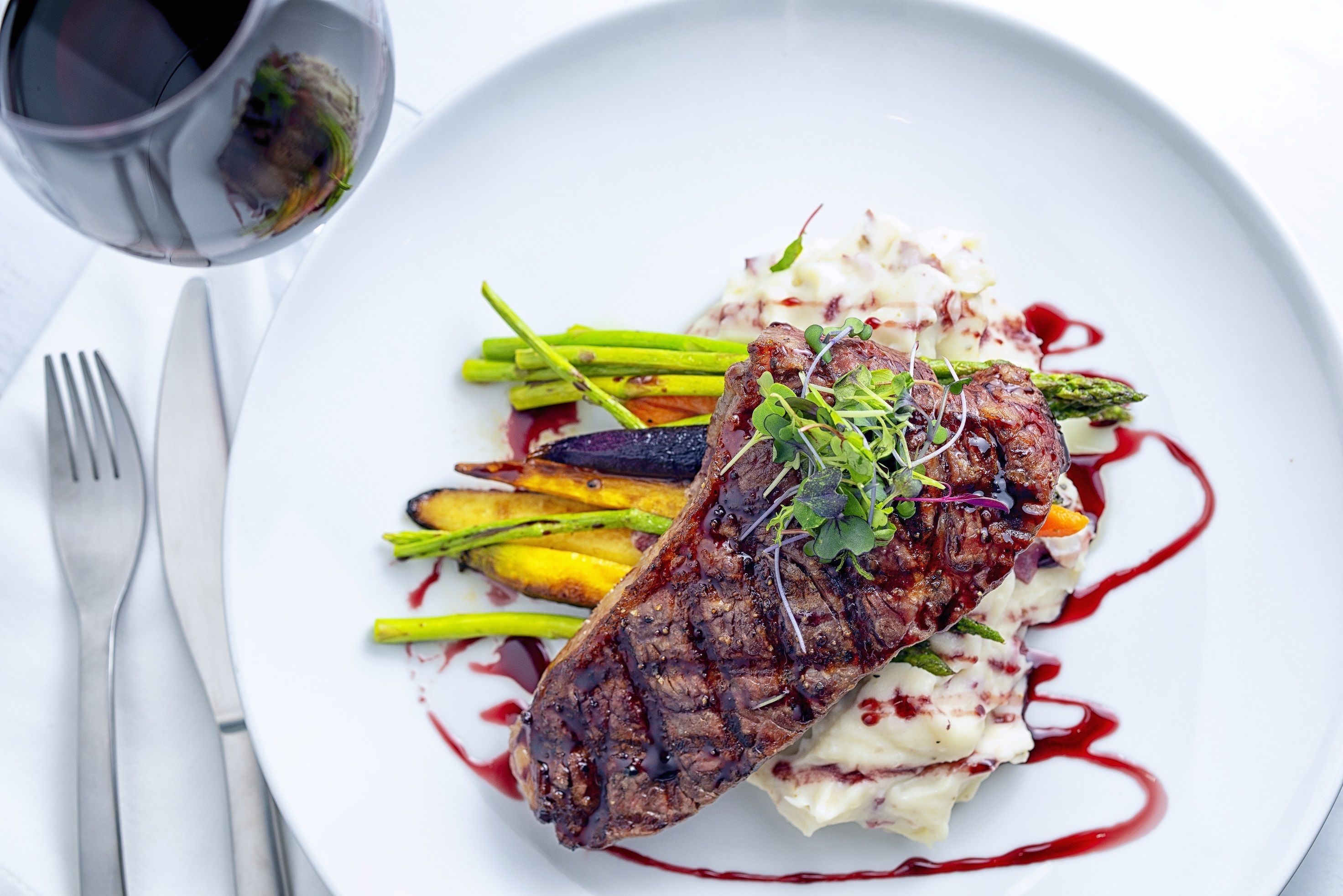 Um prato com bife, aspargos e purê de batatas, acompanhado de um copo de vinho tinto.