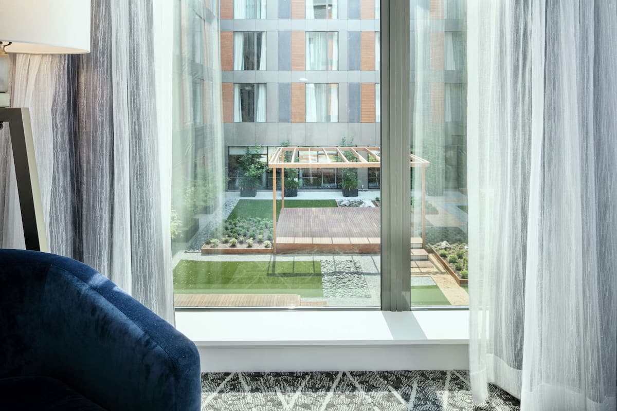 A modern room with a large window, a blue chair, and a view of a courtyard with plants and a wooden deck.