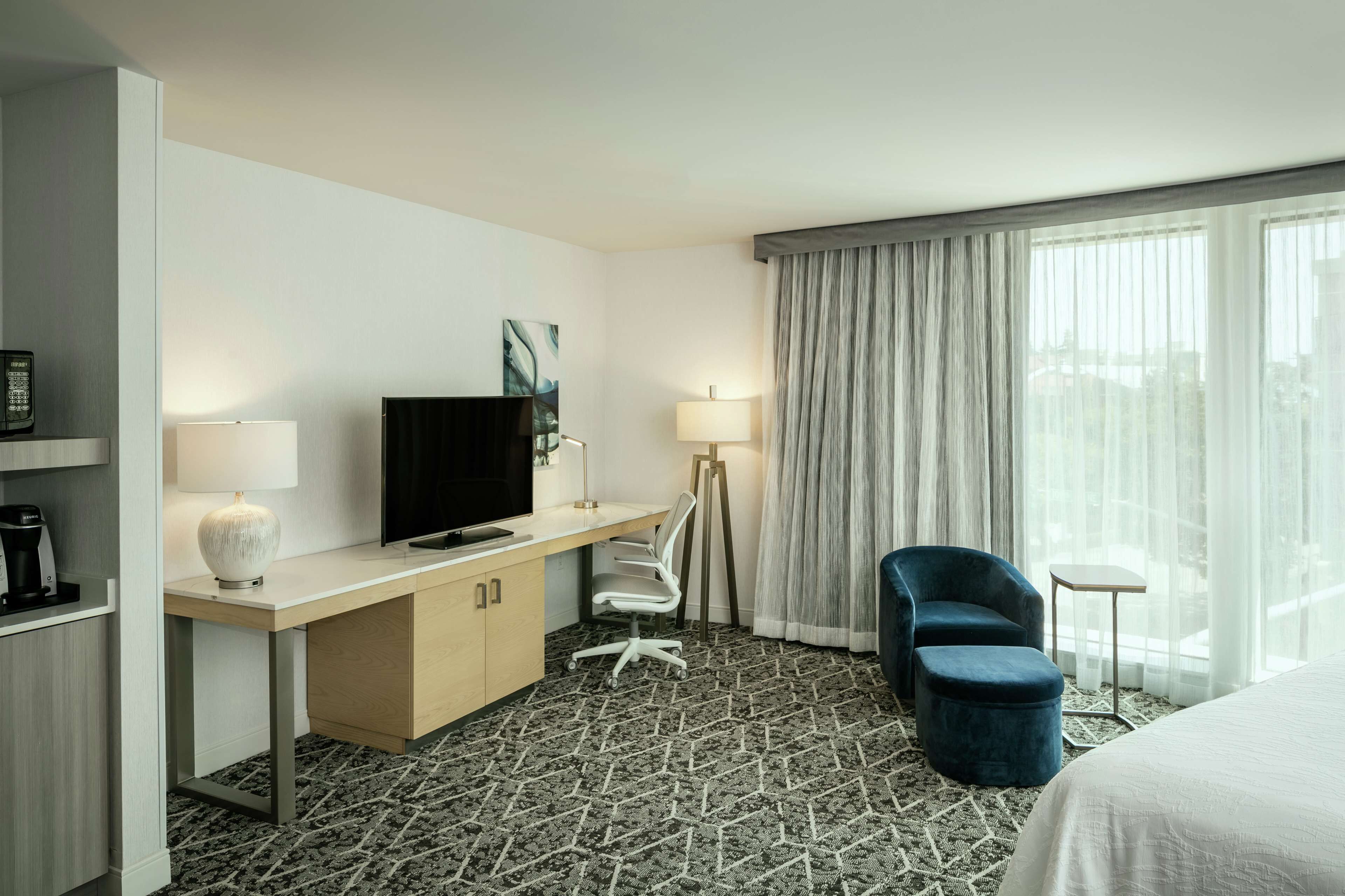 A hotel room with a bed, a desk with a TV, a chair, a lamp, and a view of the city through large windows.
