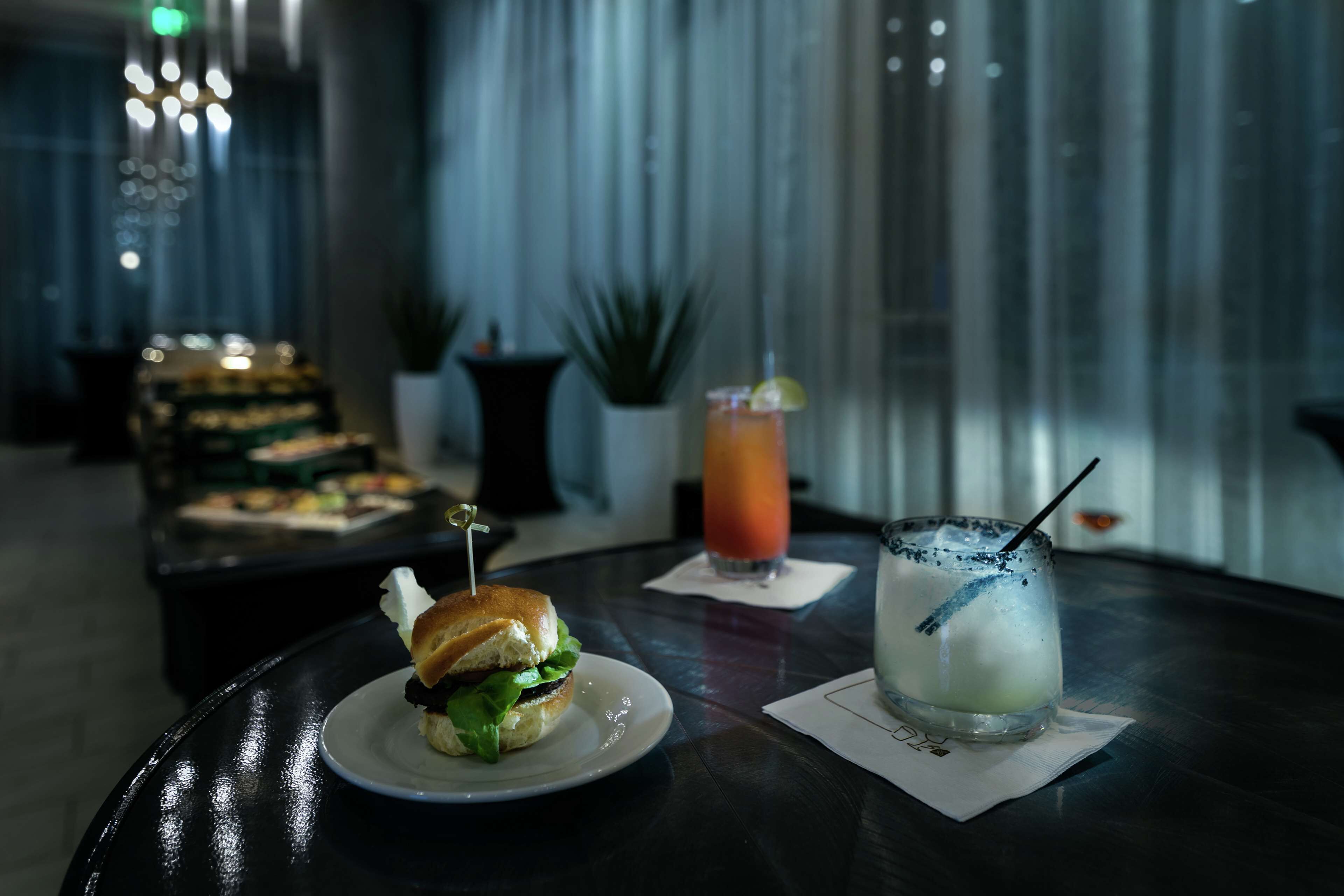 A table with a sandwich and two drinks, a plant, and a curtain in the background.