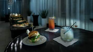 A table with a sandwich and two drinks, a plant, and a curtain in the background.