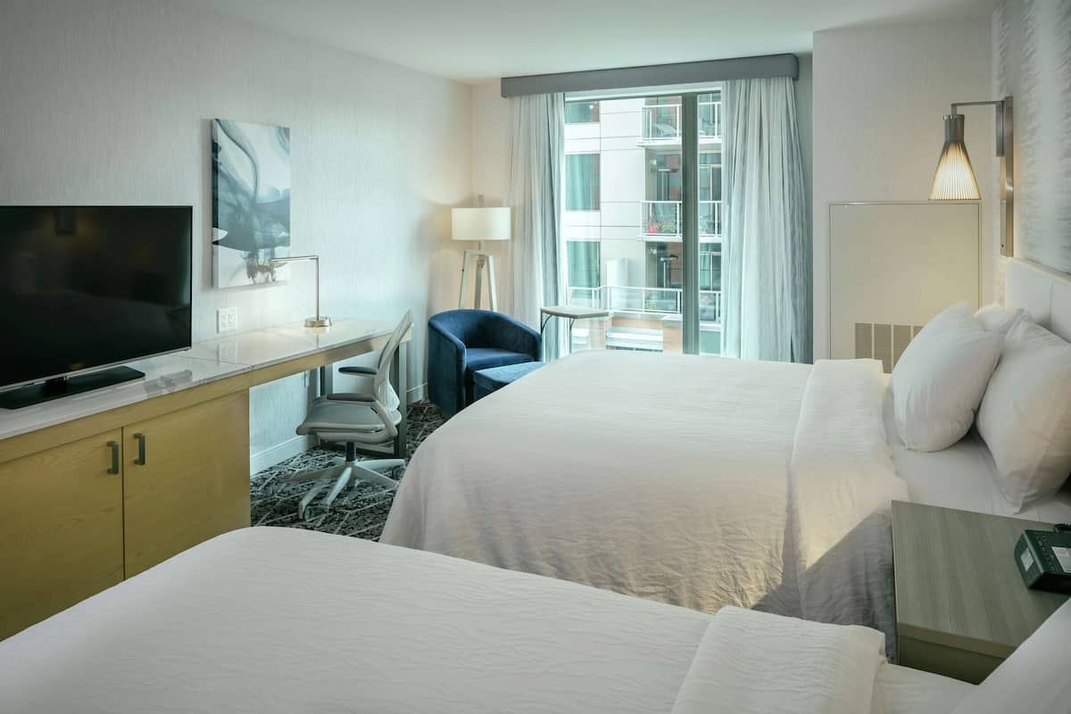A hotel room with two beds, a TV, a desk with a chair, a lamp, and a view of the outside balcony.