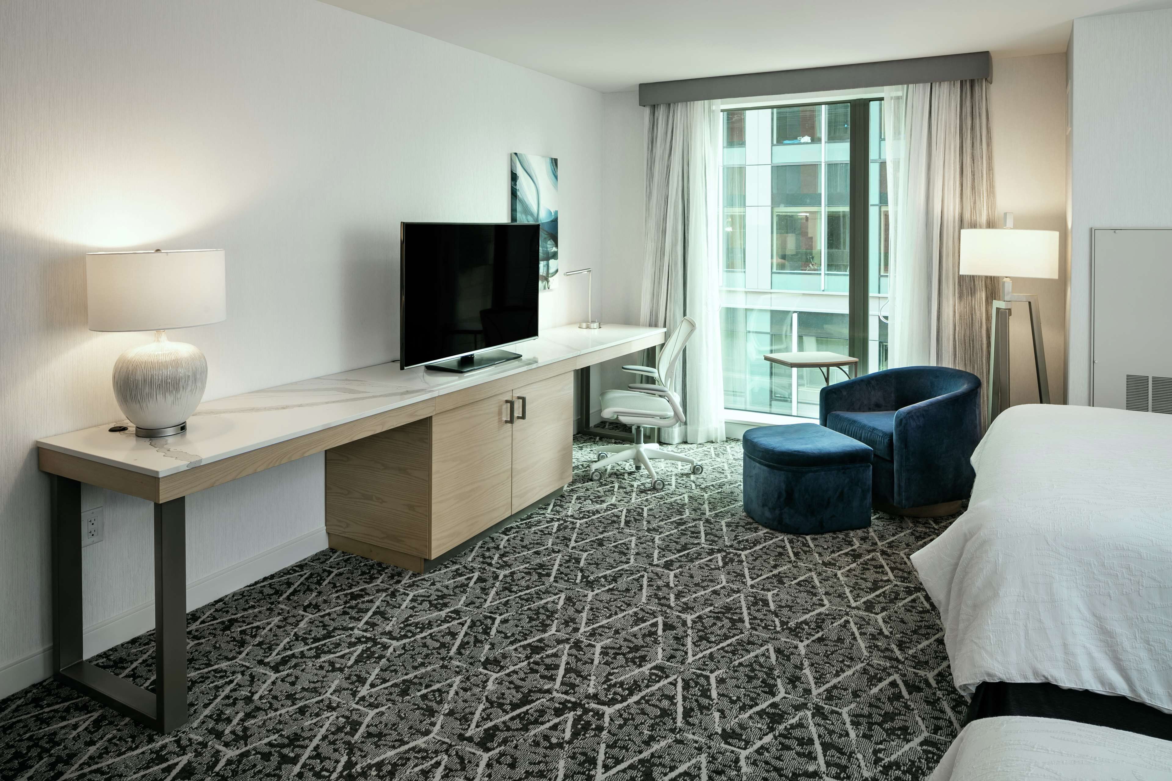 A hotel room with a desk, TV, chair, ottoman, and bed.