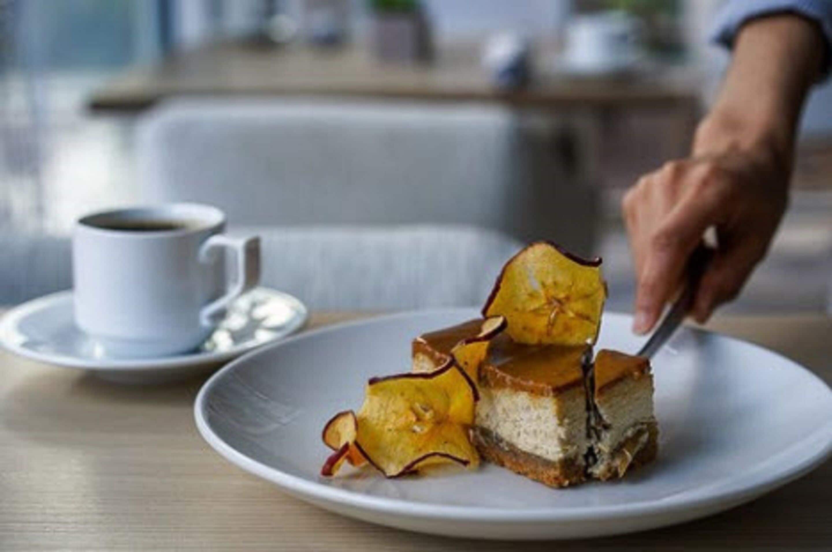 A dessert plate with a slice of cake and caramelized fruit on a white plate, a cup of coffee on a saucer, and a hand cutting the cake.