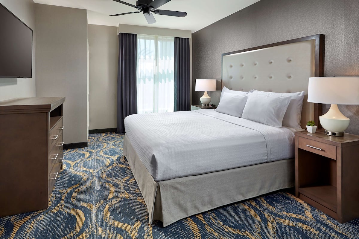 A hotel room with a large bed, two bedside tables with lamps, a ceiling fan, a TV mounted on the wall, and a patterned carpet.