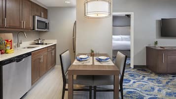Una cocina compacta con electrodomésticos modernos, un comedor con una mesa lista para dos personas, y un dormitorio visible a través de un umbral abierto.