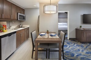 Una cocina compacta con electrodomésticos modernos, un comedor con una mesa lista para dos personas, y un dormitorio visible a través de un umbral abierto.