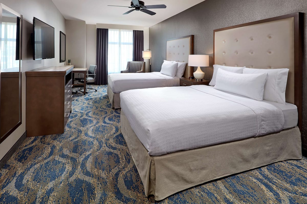 A hotel room with two beds, a desk, a chair, a TV, and a ceiling fan.