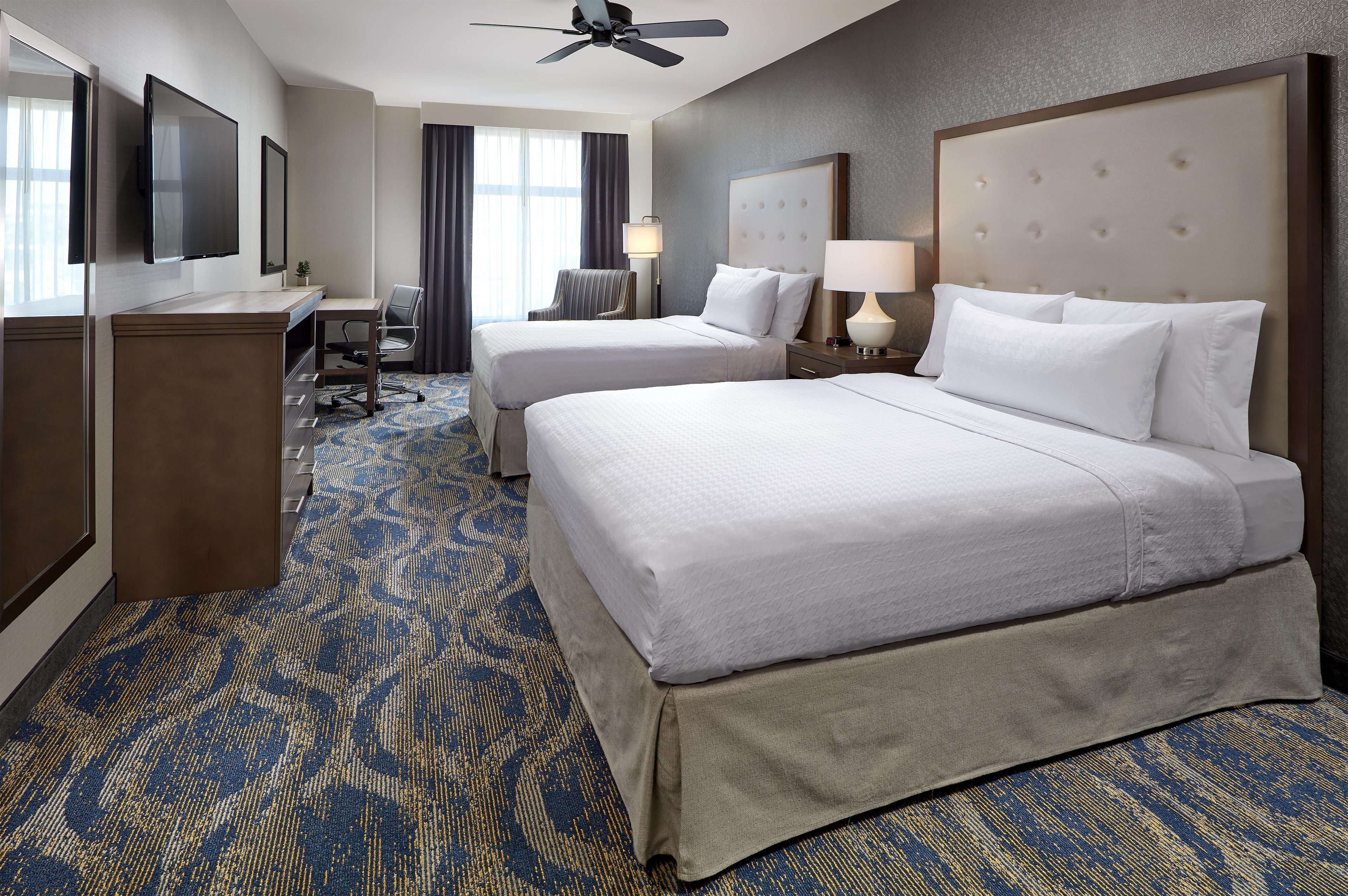 A hotel room with two beds, a desk, a chair, a TV, and a ceiling fan.