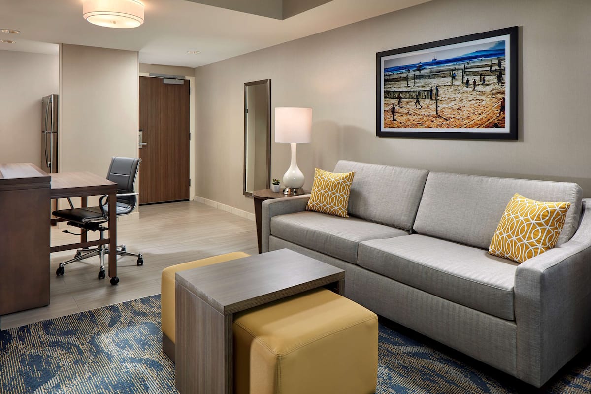 A hotel room with a grey sofa, a small table, a desk with a chair, a lamp, and a framed beach picture on the wall.