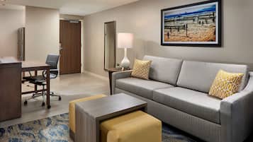 A hotel room with a grey sofa, a small table, a desk with a chair, a lamp, and a framed beach picture on the wall.