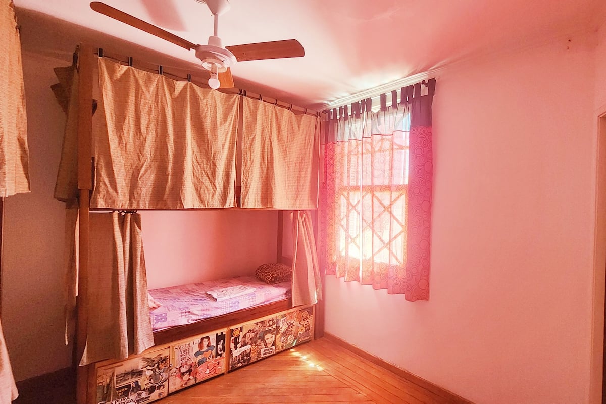 A room with a bunk bed, a ceiling fan, a window with patterned curtains, and a door.