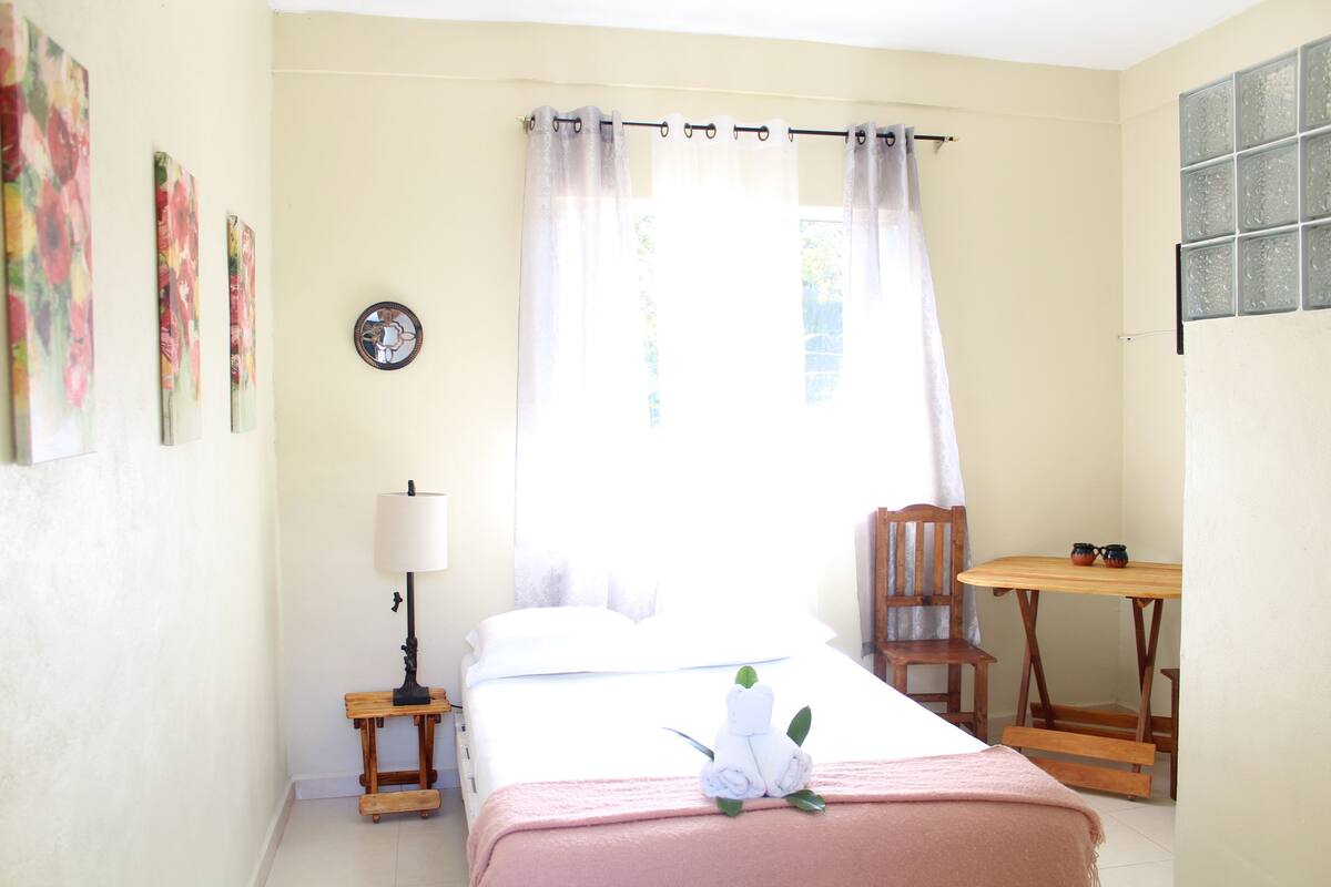 Una habitación de hotel con una cama, una mesita, una silla, una lámpara y un ventanal con cortinas.