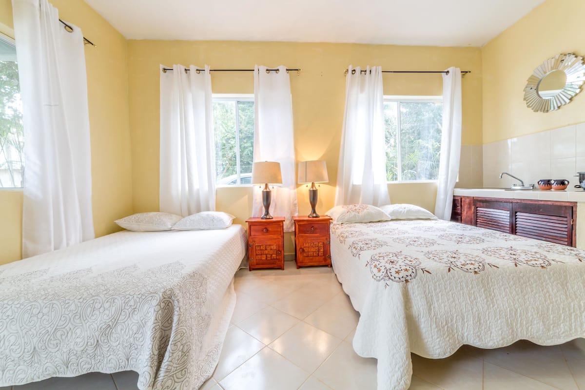 Un dormitorio con dos camas, una mesita de noche de madera con lámparas, un baño con lavamanos y espejo, y ventanas amplias con cortinas.