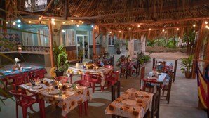 An outdoor dining area with tables and chairs, surrounded by plants and a thatched roof.
