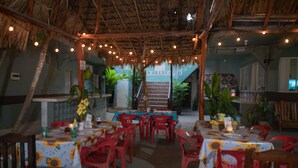 A restaurant with tables set for dining, red plastic chairs, and a thatched roof.