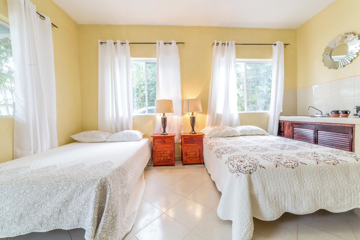 A bedroom with two beds, a wooden nightstand with lamps, a bathroom with a sink and mirror, and large windows with curtains.