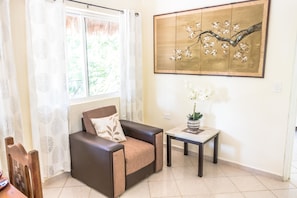 A living room with a sofa, a small table with a potted plant, a window with curtains, and a wall-mounted artwork. - Mayan Villas Hotel (Chiquilá)