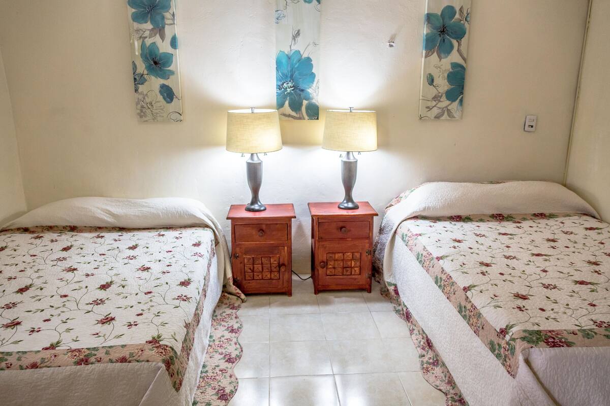 Un dormitorio con dos camas, cada una con una colcha floral, flanqueadas por mesitas de noche de madera con lámparas.