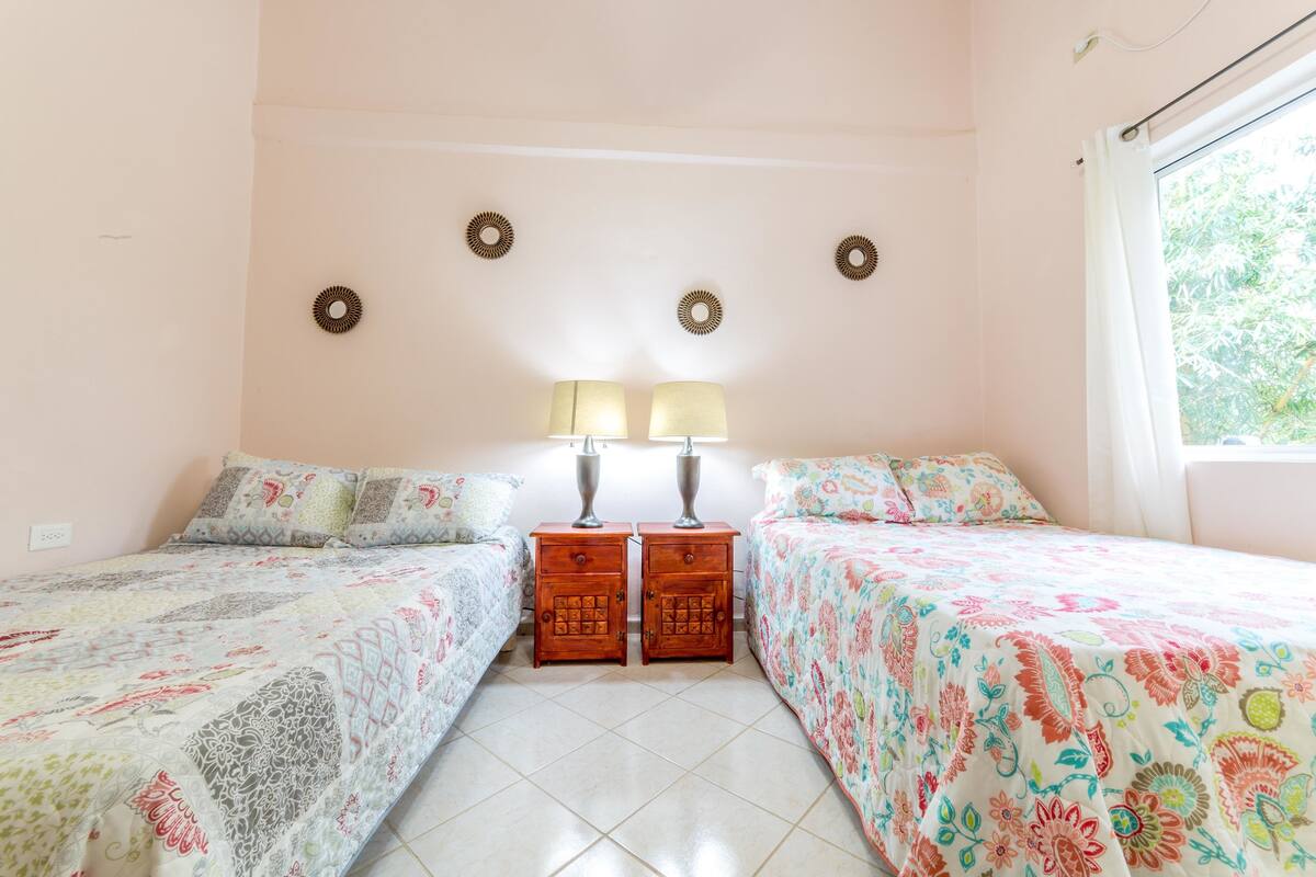 Un dormitorio con dos camas, una mesita de noche con lámparas y una ventana con cortinas.