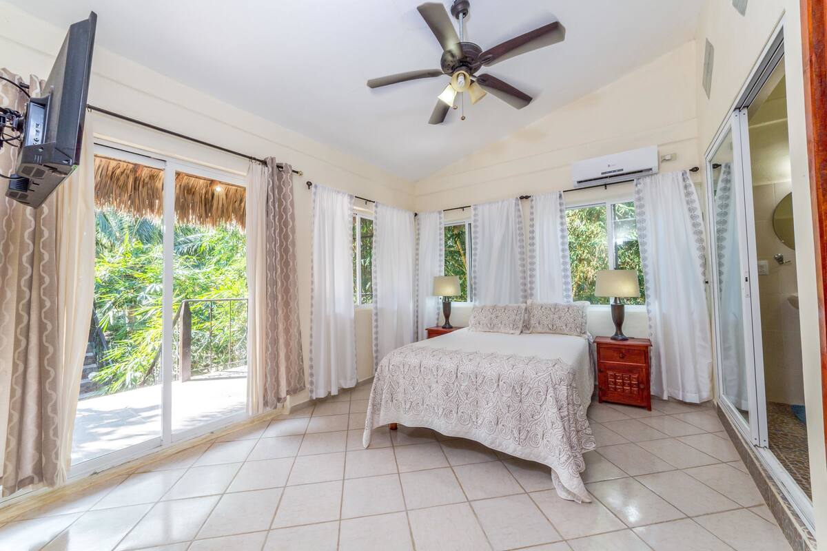 Un dormitorio con cama, ventilador de techo, televisor y ventana con cortinas.